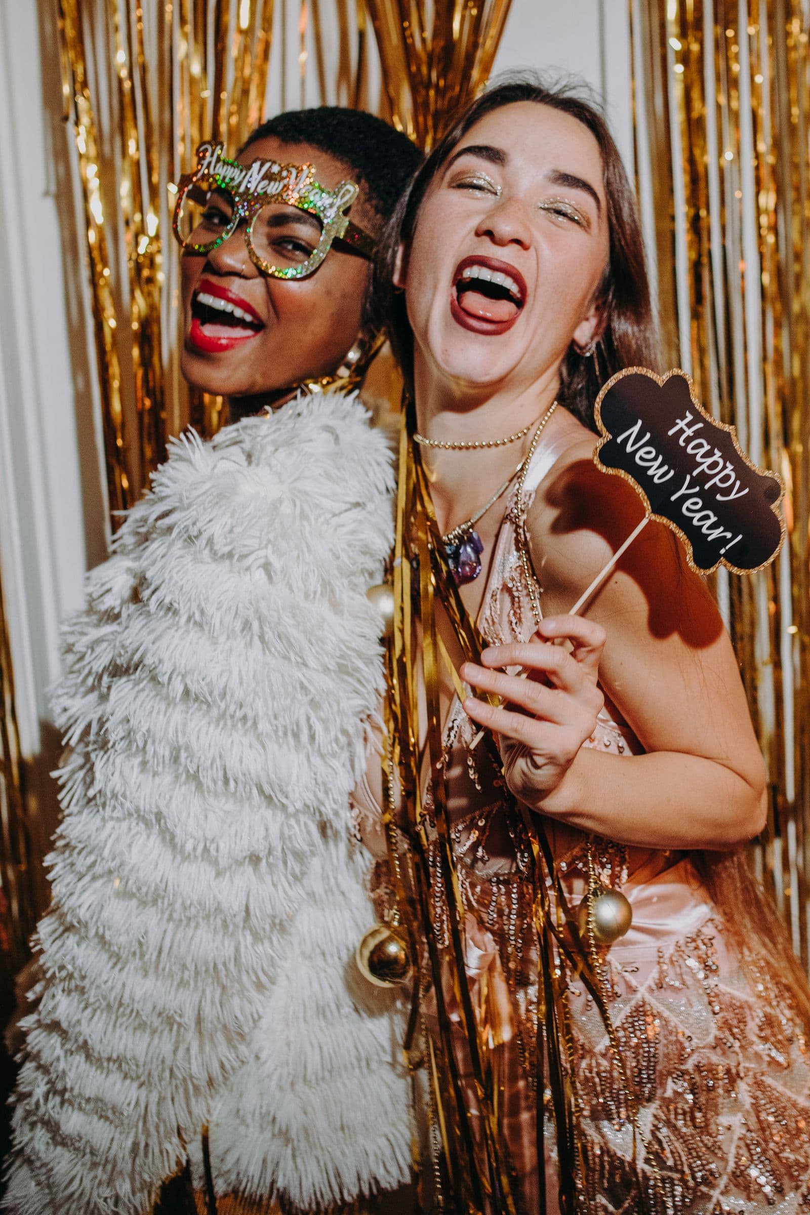 Two guests laughing with party props in front of a gold tinsel backdrop